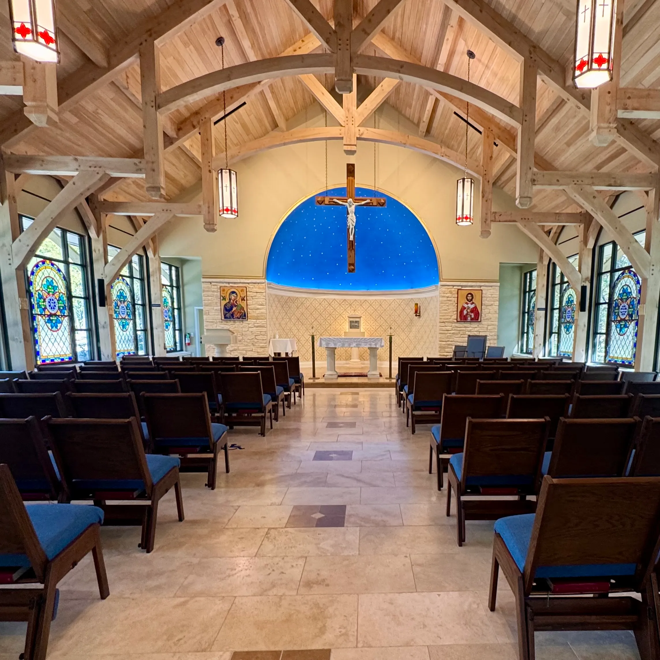 The interior of a chapel with a timber-framed vaulted ceiling, a blue half-dome painted with stars above a crucifix, stained-glass windows, and rows of blue-cushioned chairs.
