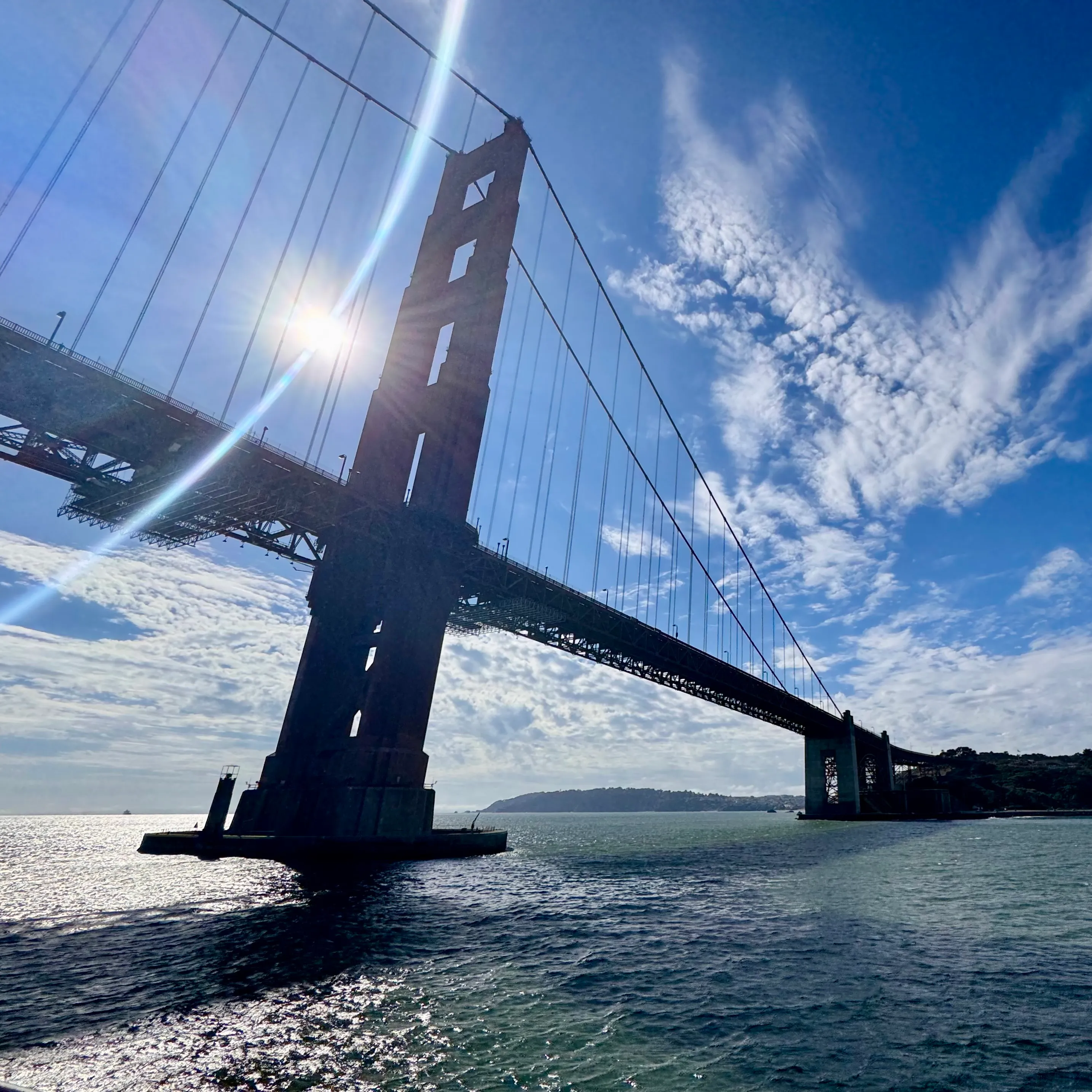 The Golden Gate Bridge seen from water level, sunlight flaring through its towers against a blue sky.