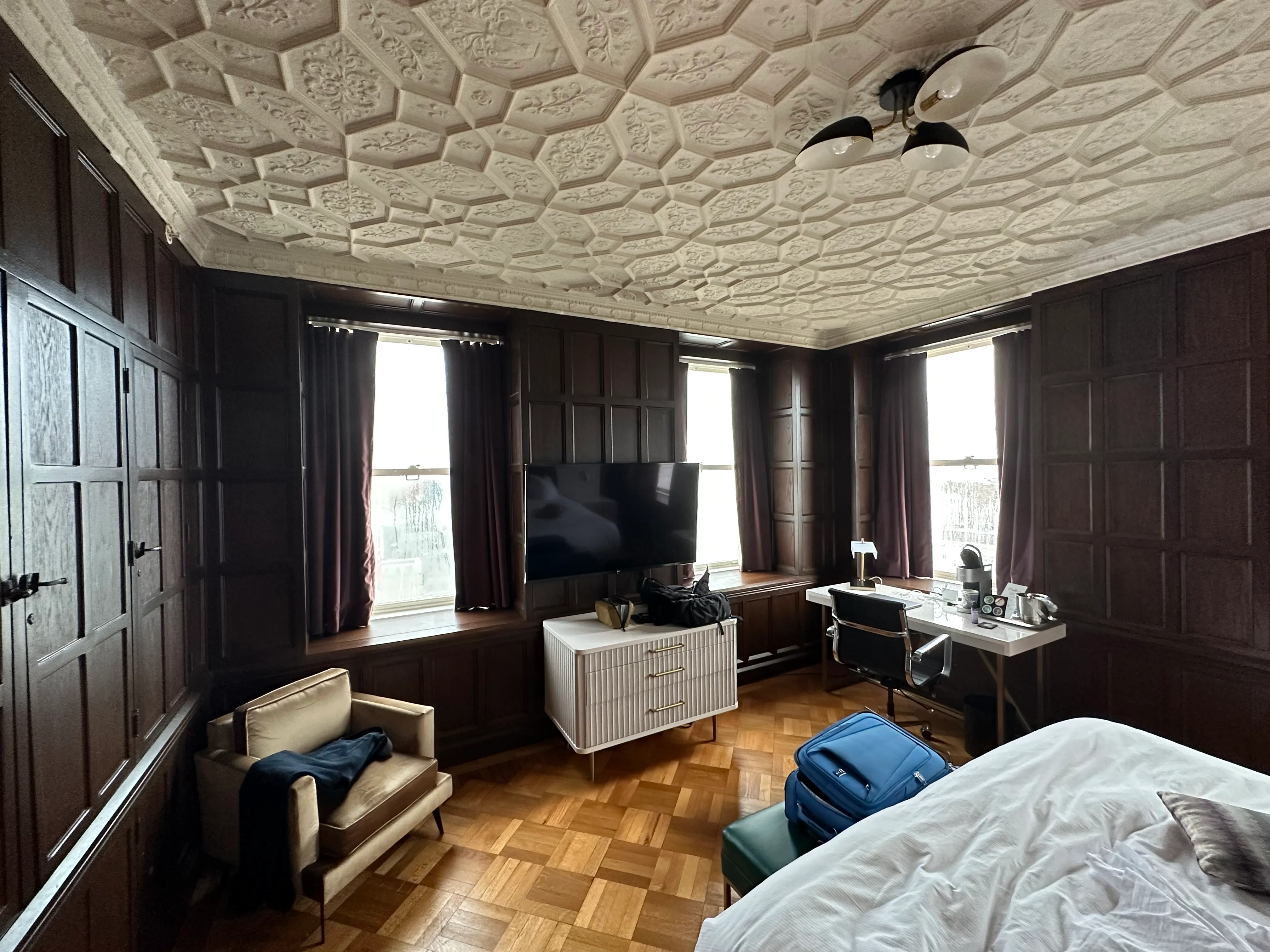 A historic hotel room with dark wood-paneled walls, an ornate plaster ceiling, and a blue suitcase resting on the bed.