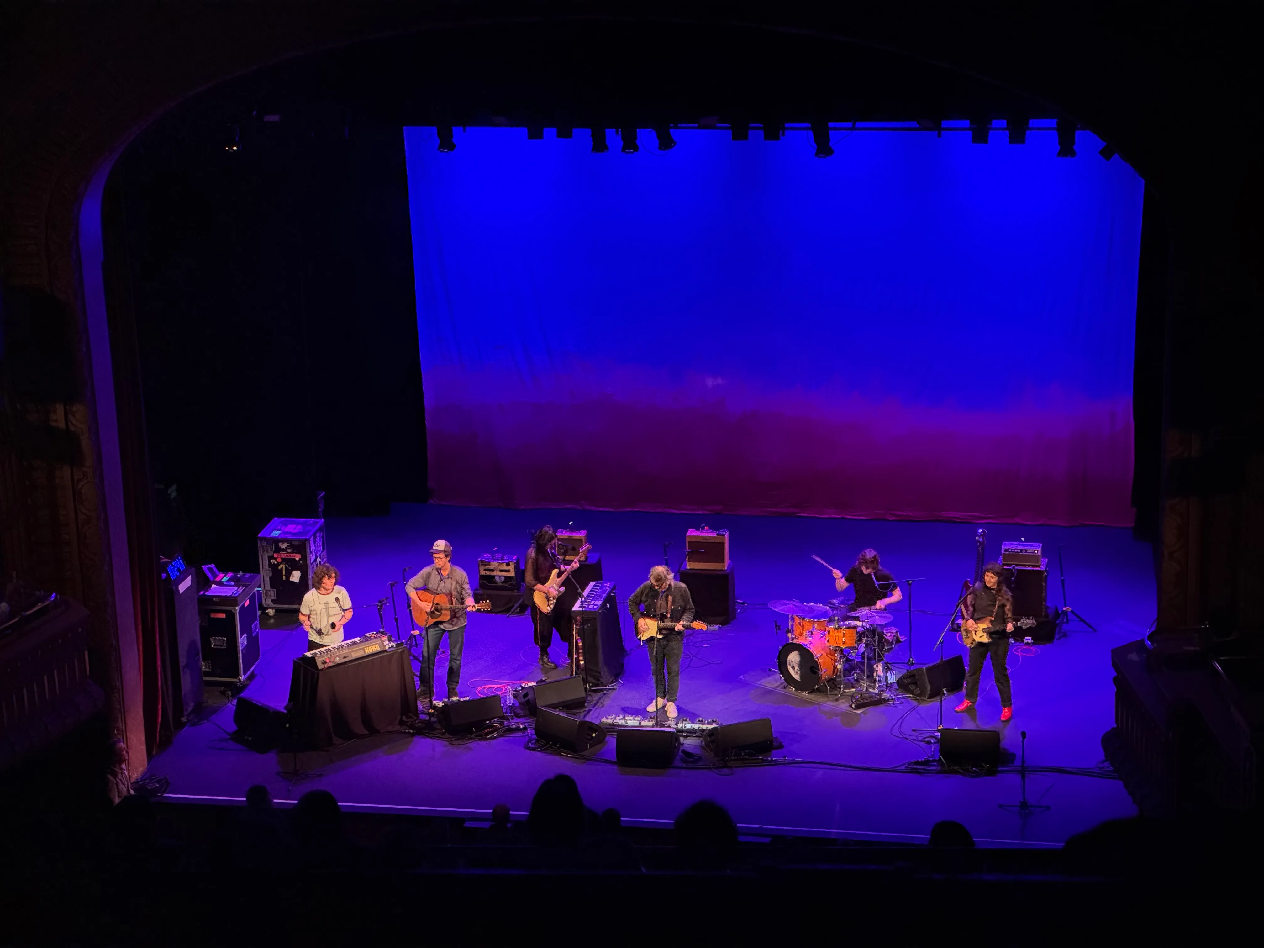 A six-piece band performing on a theater stage under deep blue light.