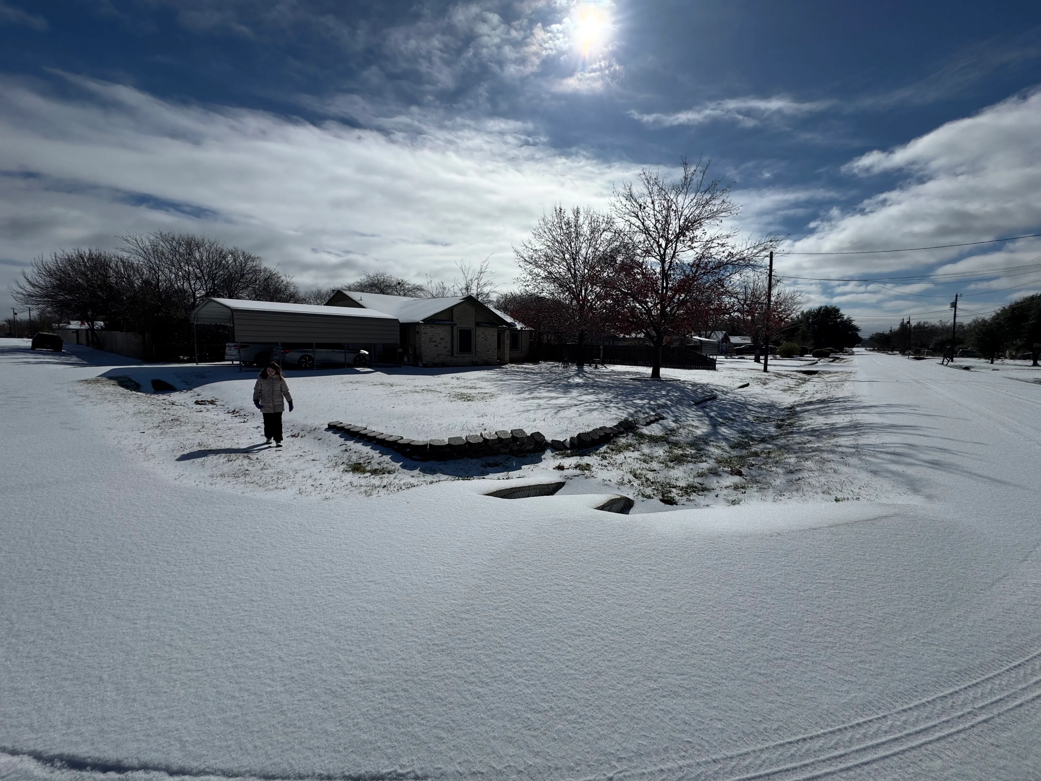 A child in a puffy coat walks across fresh snow in front of a single-story brick house under a bright blue sky.